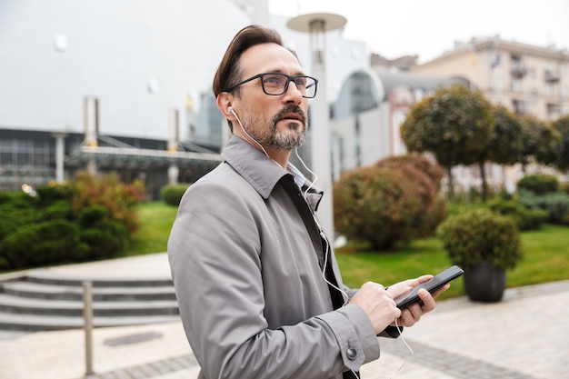 Image of handsome businessman in eyeglasses using cellphone and earphones while walking at city street