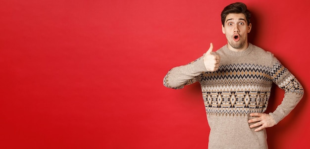 Image of handsome caucasian man in christmas sweater looking amazed showing thumbup in approval like something standing over red background