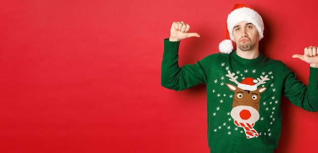 Image of handsome and confident young man in green sweater and santa hat pointing at himself celebrating christmas standing over red background