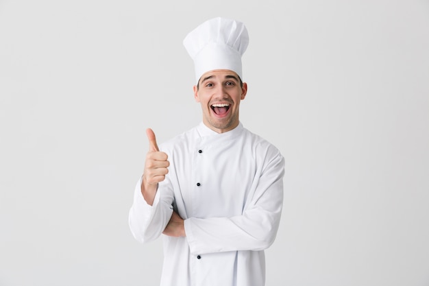 Image of handsome excited young man chef indoors isolated over white wall background showing thumbs up gesture.