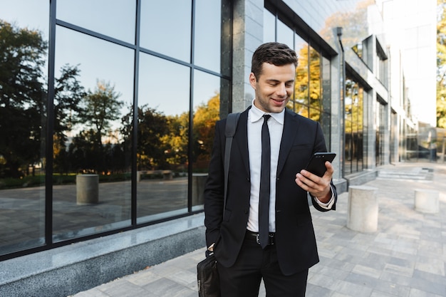 Image of a handsome happy young businessman walking outdoors near business center using mobile phone.