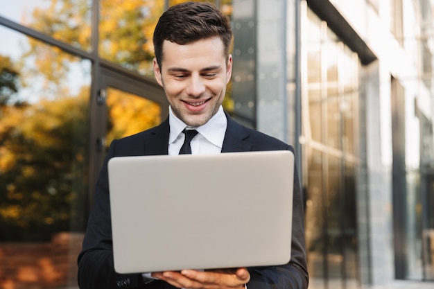 Image of a handsome happy young businessman walking outdoors using laptop computer.