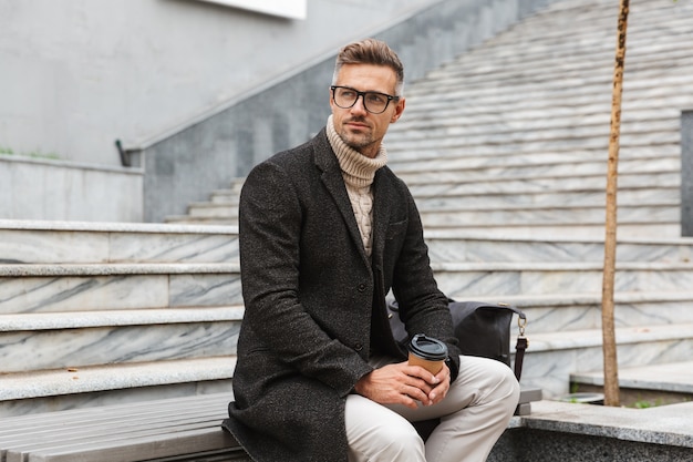 Image of handsome man 30s wearing eyeglasses, sitting on stairs with takeaway coffee while strolling through city street