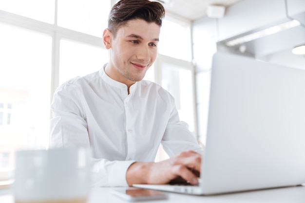 Image of handsome man dressed in white shirt sitting near cup of coffee while using laptop computer. Coworking. Looking at computer.