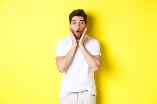 Image of handsome man looking surprised, gasping and saying wow, looking at promo offer amazed, standing over yellow background