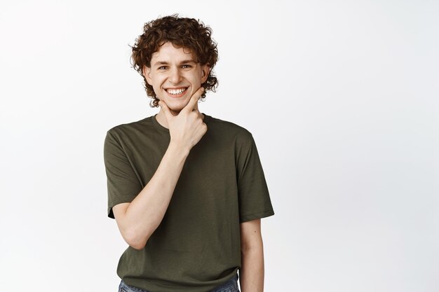 Image of handsome smiling curlyhaired guy touching his chin and looking intrigued thinking of smth standing over white background