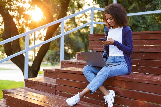 Image of happy african young woman sitting outdoors in park on steps using laptop computer and mobile phone.