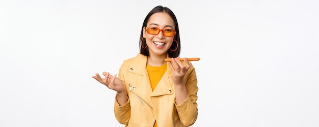 Image of happy asian girl talking on speakerphone recording translating her voice with mobile phone app talking in smartphone dynamic standing over white background