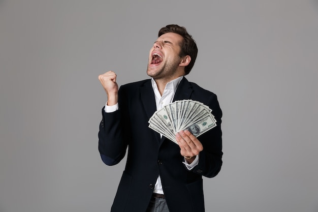 Image of happy businessman 30s in suit smiling and holding fan of money in dollar banknotes, isolated over gray wall