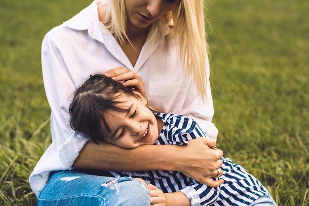 Image of happy cute little girl embrace and lying on her mother legs outdoor Loving mother and daughter sitting on green grass in the park Mom and kid cuddling have fun outside Good relationship