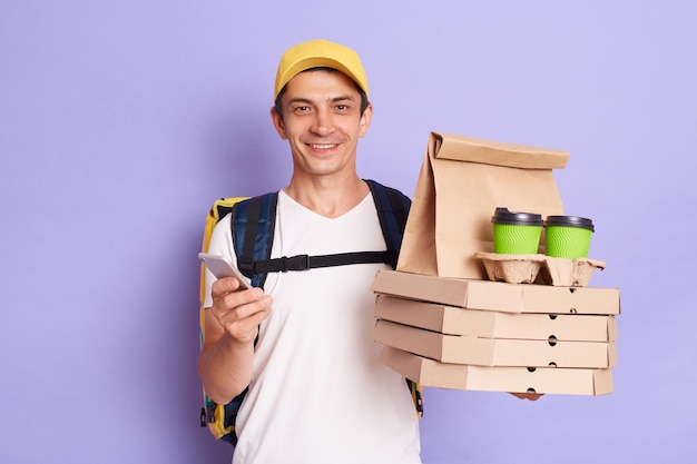 Image of happy delivery man in cap and white Tshirt using smart phone holding pizza boxes and coffee cheking address for handing parcel isolated on purple background