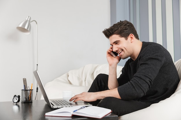 Image of a happy handsome young man at home indoors using laptop computer talking by mobile phone.