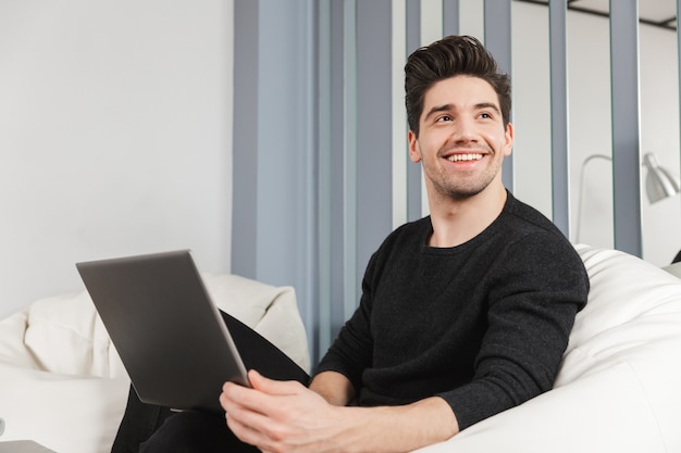 Image of a happy handsome young man at home indoors using laptop computer.