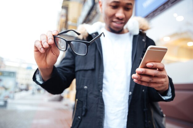 Image of happy man holding his eyeglasses and phone in hands while walking outdoors. Look at phone.