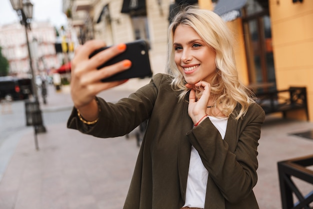 Image of happy woman smiling and taking selfie photo, while walking through city street