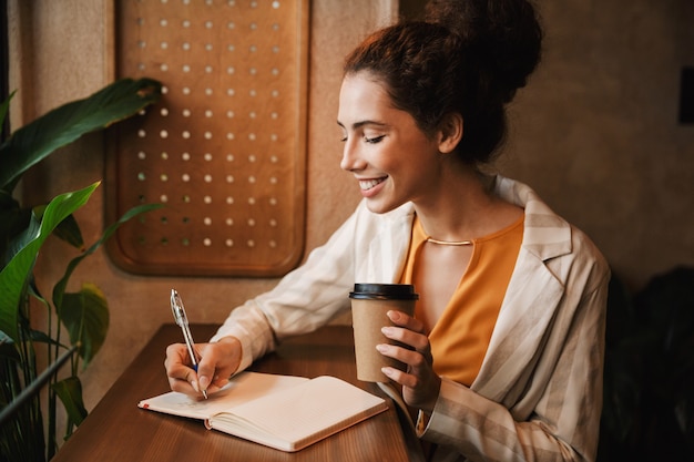 Image of a happy young business woman indoors in cafe drinking coffee writing notes.