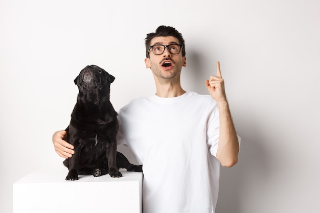 Image of happy young man and dog both looking up at promo, owner pointing finger at top, pug staring at loog, standing over white background