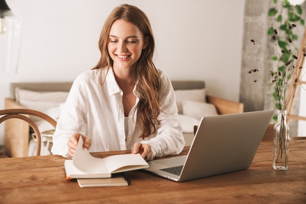 Image of a happy young pretty woman sit indoors in office using laptop computer holding notebook.