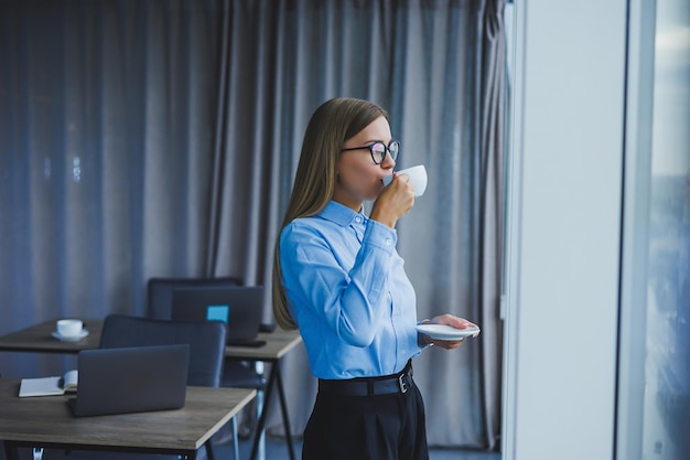 Image of a happy young woman in a classic shirt and glasses smiling and drinking coffee while standing by the window in a modern office with large windows Remote work