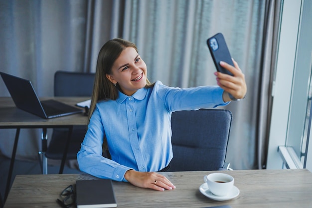 Image of a happy young woman in a jacket smiling and working on a laptop talking on the phone in a modern office with large windows Remote work