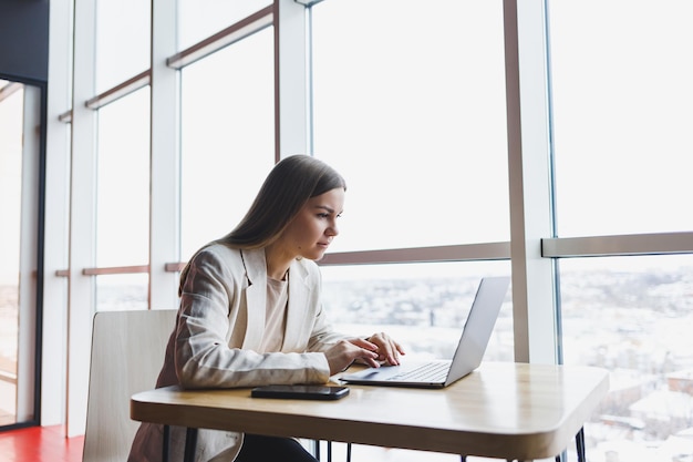 Image of a happy young woman in a jacket smiling and working on a laptop while talking on the phone in a modern office with large windows Remote work