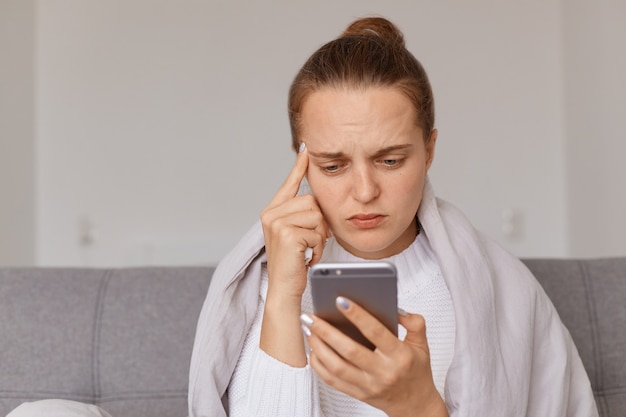 Image of ill woman with hair bun sitting on sofa wrapped in blanket, having high temperature and terrible headache, keeping finger on her temple, dialing doctor's number.