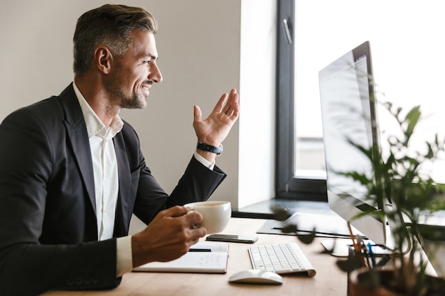 Image of joyful businessman 30s wearing suit drinking coffee while working on computer in office