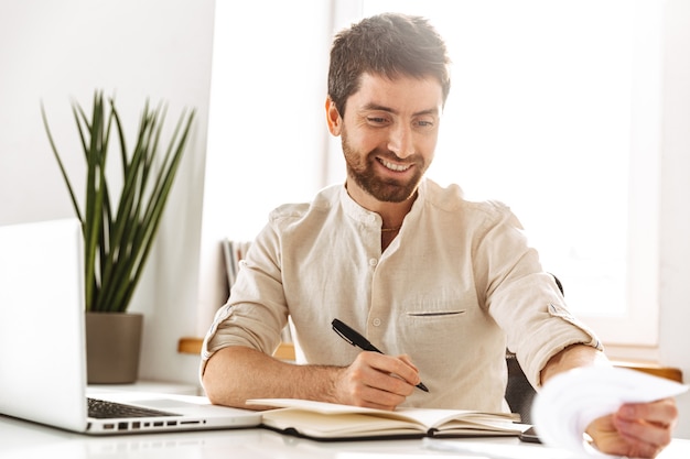 Image of joyful businessman 30s wearing white shirt working with laptop and paper documents, while sitting in bright office