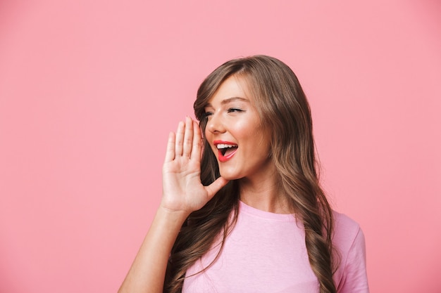 Image of joyful european woman with long curly brown hair looking aside at copyspace and shouting or calling someone, isolated over pink background