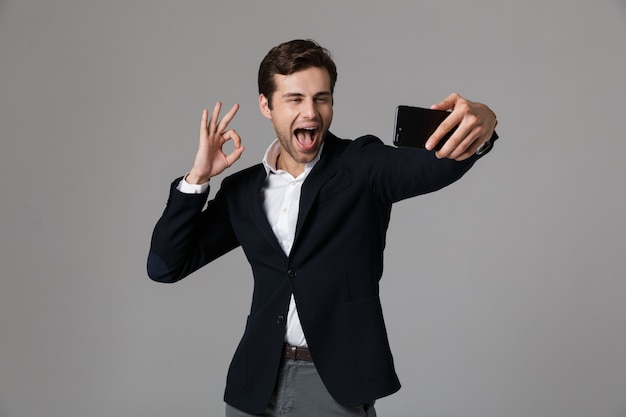 Image of joyous businessman 30s in formal suit showing ok sign while taking selfie photo on cell phone, isolated over gray wall