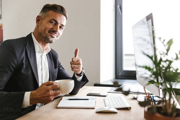 Image of joyous businessman 30s wearing suit drinking coffee while working on computer in office