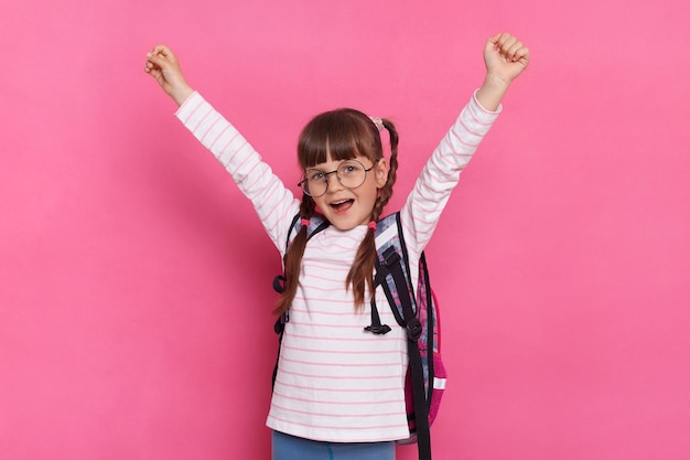 Image of little schoolgirl wearing shirt and eyeglasses standing with backpack isolated over pink background raised arms screaming with happy facial expression