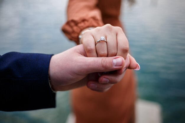 Image of a man putting a silver wedding ring on a woman's hand, outdoors. Sea or river background