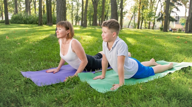 Image of middle aged woman teaching teenage boy doing yoga and fitness on grass at park. Family taking care of their health