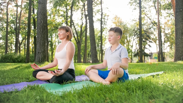 Image of middle aged woman teaching teenage boy doing yoga and fitness on grass at park. Family taking care of their health