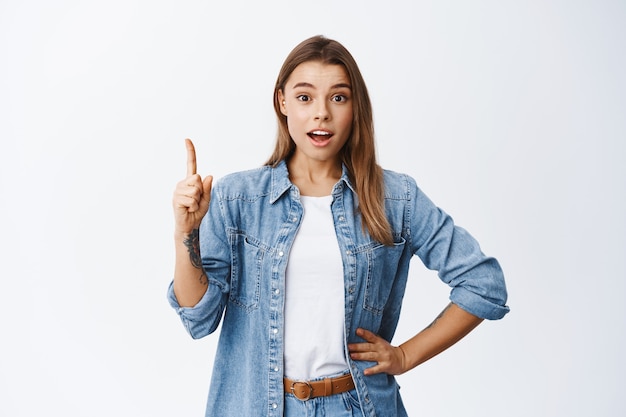 Image of millennial girl raising finger up in eureka gesture, pitching an idea, pointing finger up to say suggestion, recommending an advertisement, white wall