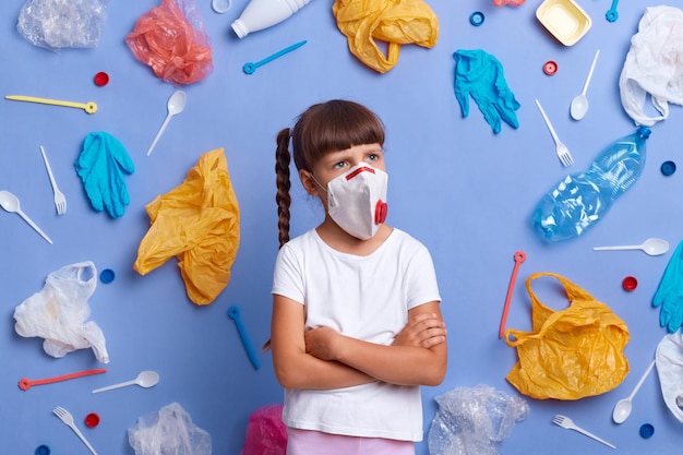 Image of pensive thoughtful little girl wearing white t shirt and protective respirator posing against blue wall and much plastic garbage around thinking about air pollution and Earth protection