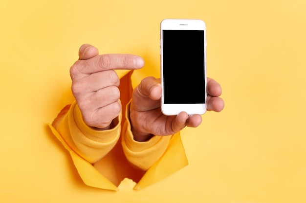 Image of person hand breaking through yellow paper and holding smart phone pointing at blank screen with copy space for advertisement or promotional text