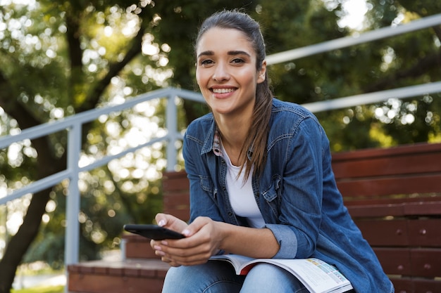 Image of pretty happy cheerful young caucasian woman sitting outdoors chatting by mobile phone.