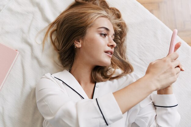 Image of pretty young girl wearing white pajama holding cellphone while lying in bed at home