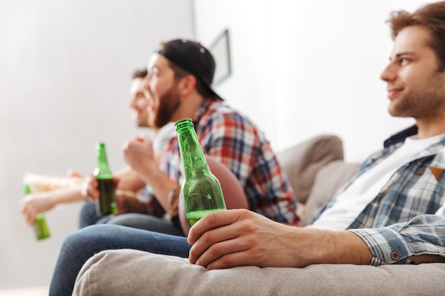 Image in profile of young bachelors 30s resting at home, and watching football game on tv with drinking beer