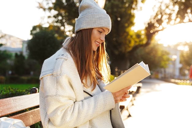 Image of a redhead woman sitting on bench outdoors holding reading book.