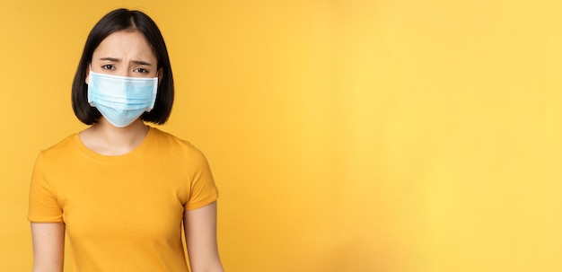 Image of sad and disappointed asian woman in medical mask frowning and looking displeased standing against yellow background