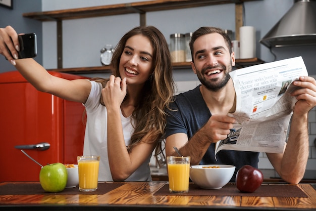 Image of satisfied couple man and woman taking selfie photo on cell phone while having breakfast in kitchen at home