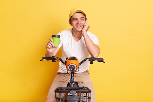 Image of satisfied man wearing white t shirt and cap riding electric scooter and stops to drink takeaway coffee enjoying his weekend isolated on yellow background