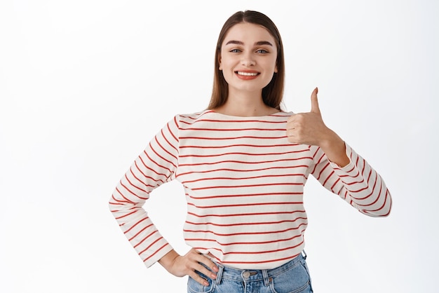 Image of satisfied young female client shows thumbs up in approval leave positive feedback pleased with good choice praise nice work make well done gesture standing over white background