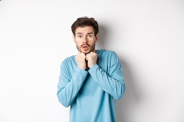 Image of scared young man trembling from fear, holding hands near face and staring at camera startled, standing over white background