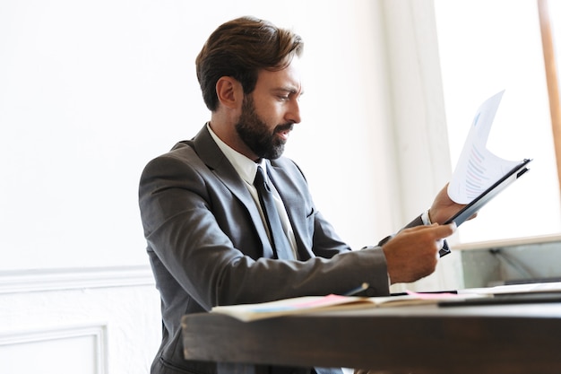 Image of serious focused businessman wearing formal suit reading documents while working in office