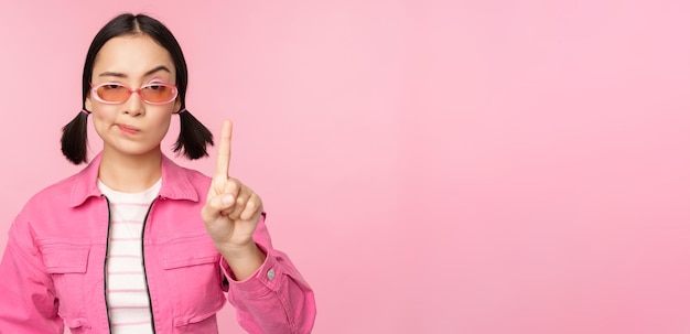 Image of serious stylish asian girl in sunglasses showing stop prohibit gesture taboo sign saying no standing over pink background