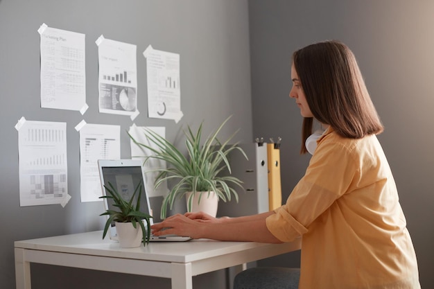 Image of serious woman freelancer wearing yellow shirt having brown hair typing on notebook in office having concentrated facile expression doing report or working on project